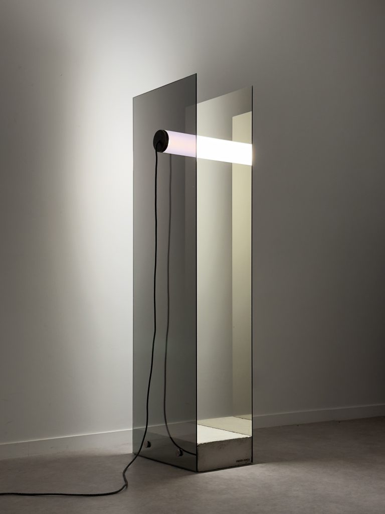 Horizontal luminous tube held by two vertical and parallel one-way mirror plates, supported by a raw concrete block.