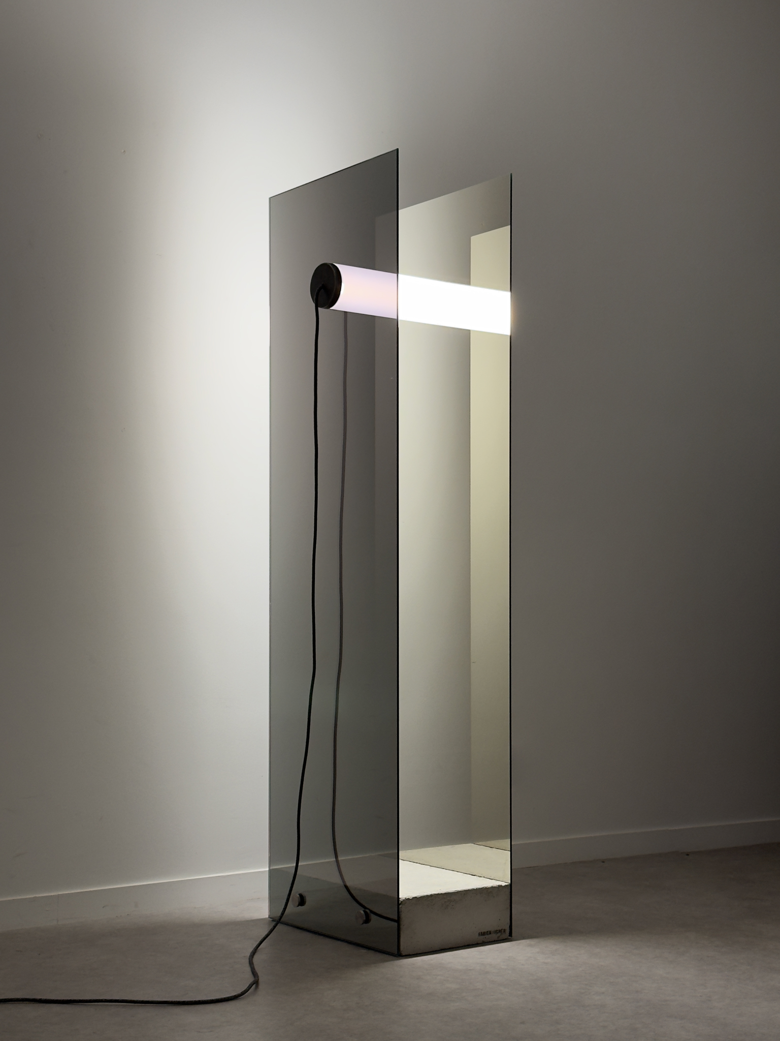 Horizontal luminous tube held by two vertical and parallel one-way mirror plates, supported by a raw concrete block.