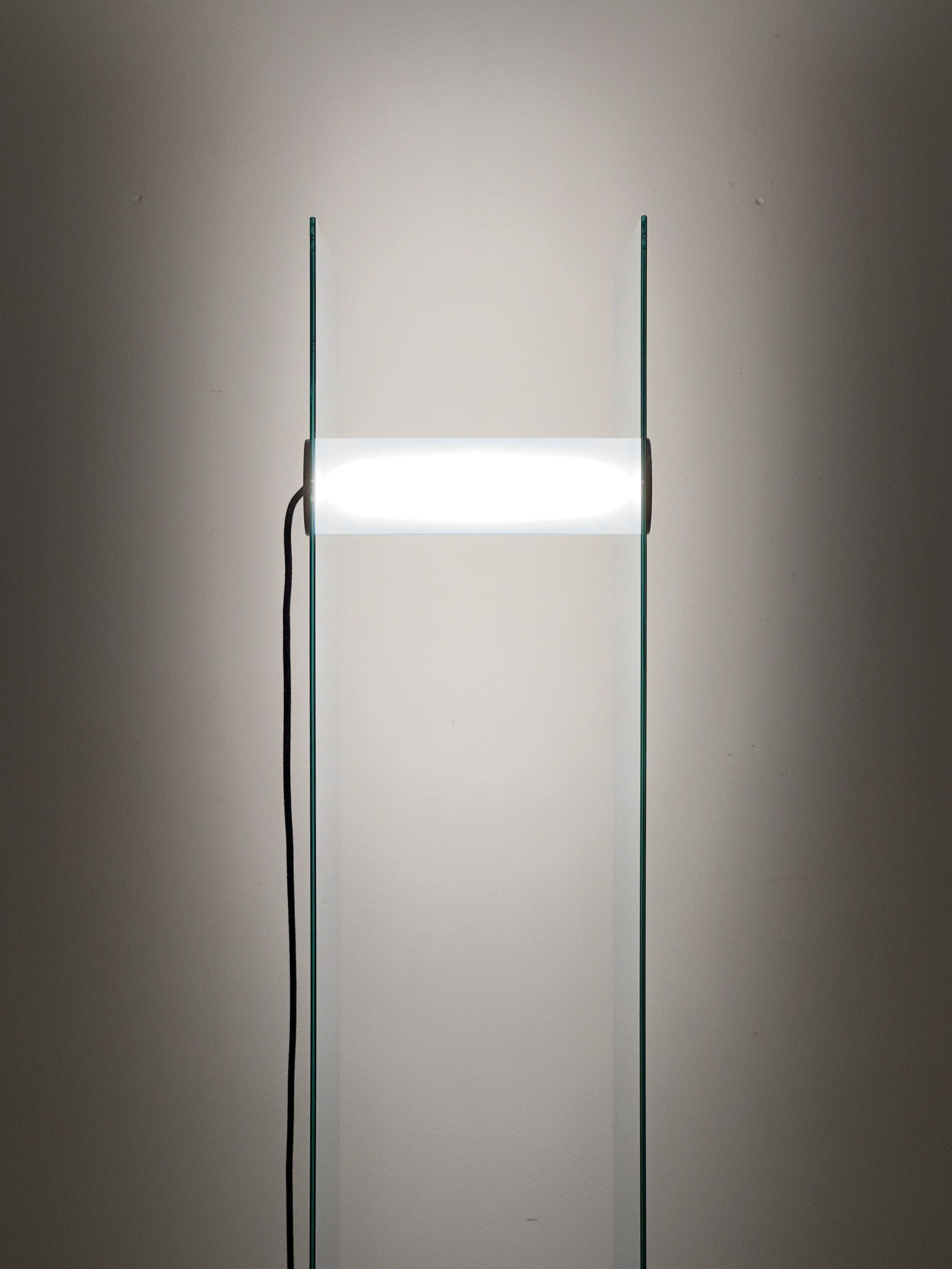 Luminous tube held between two parallel vertical mirrored tinted glass plates, with the electrical cable of the tube hanging.