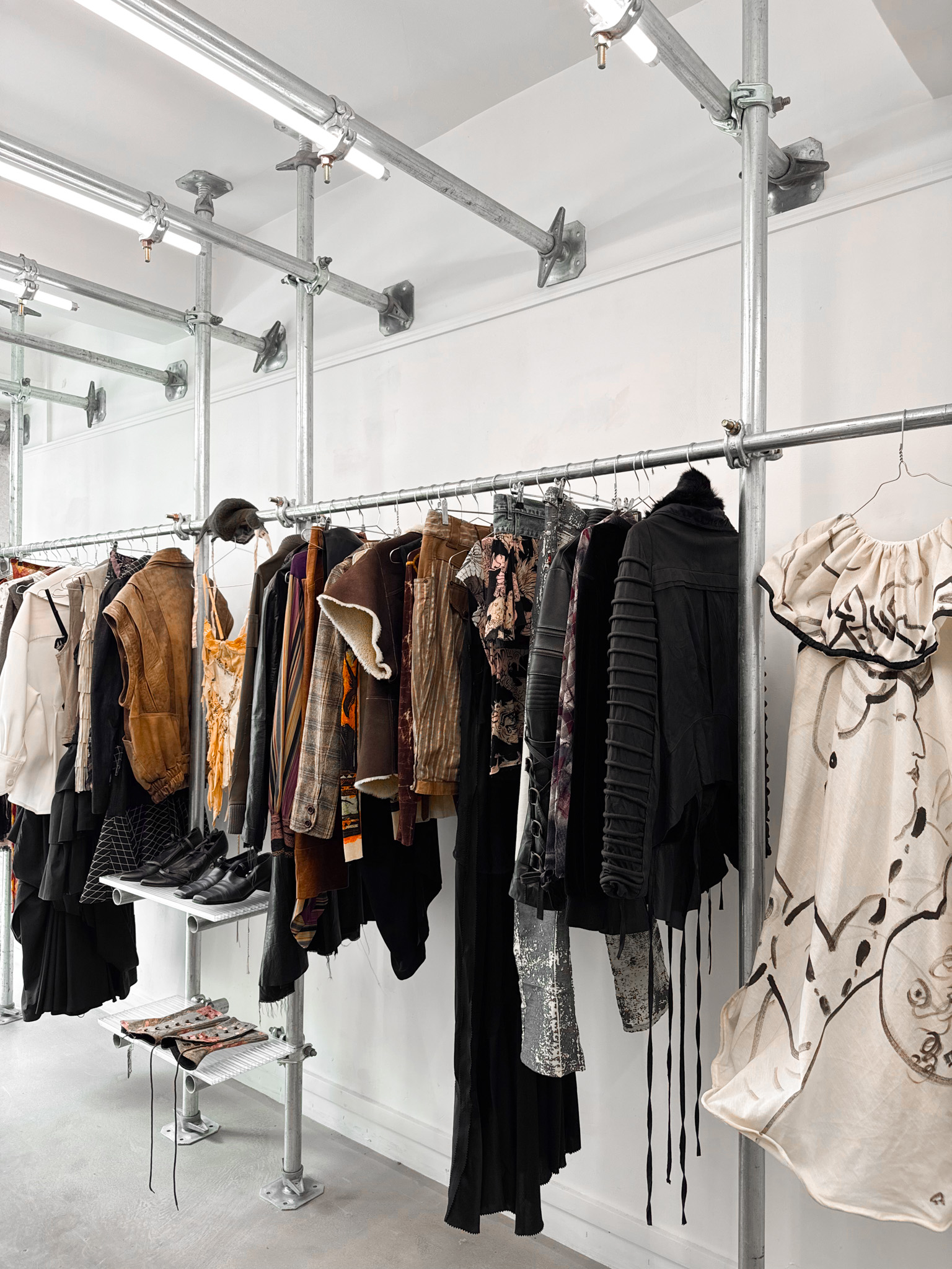 Clothes hang on galvanized steel racks in an industrial-style boutique, with shoes displayed on small metal shelves beneath bright lighting.