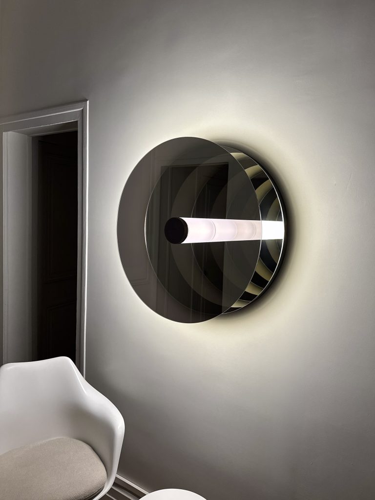 'Paramount Ragtime' is a circular wall sculpture with a glowing tube and mirrored surfaces, creating infinite reflections with the minimalist space. Positioned next to a sleek, modern chair, the sculpture interacts with the room’s architecture.