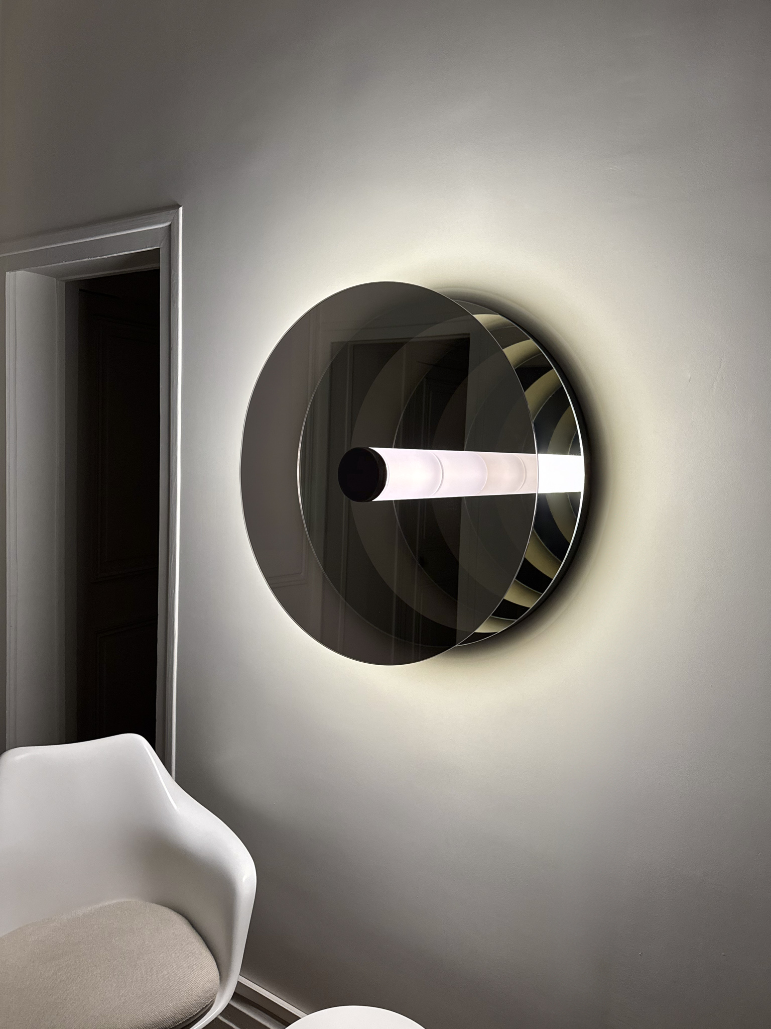 'Paramount Ragtime' is a circular wall sculpture with a glowing tube and mirrored surfaces, creating infinite reflections with the minimalist space. Positioned next to a sleek, modern chair, the sculpture interacts with the room’s architecture.