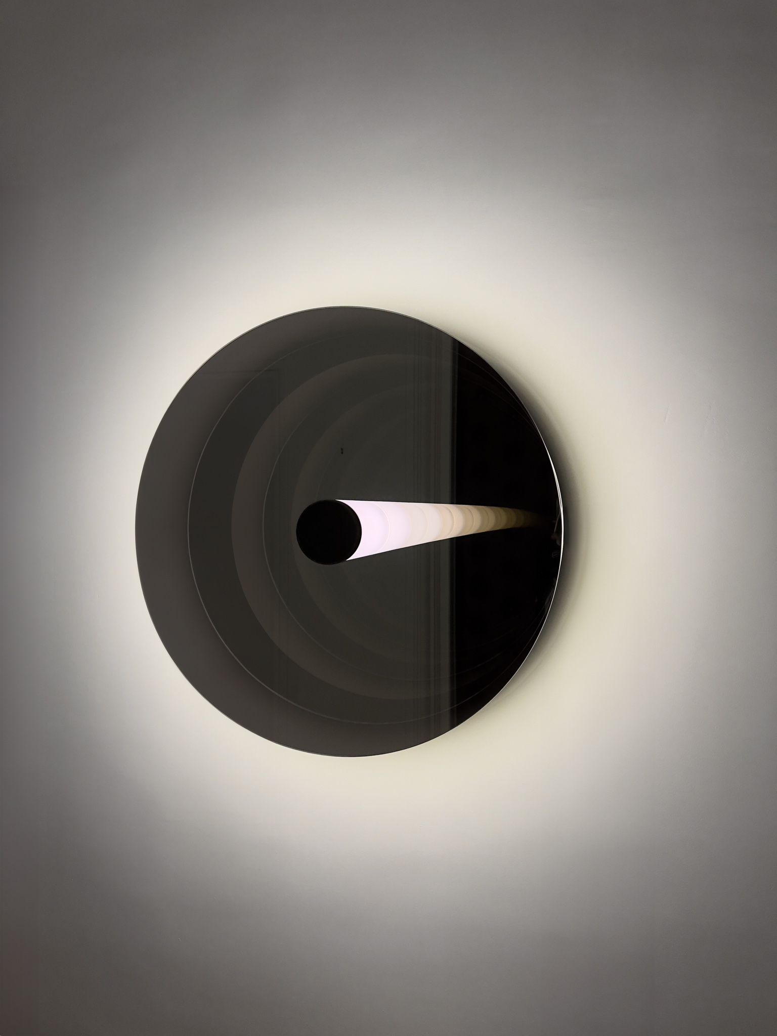 A circular wall sculpture with a luminous central tube, creating infinite reflections and soft light, evoking fluidity and timelessness.